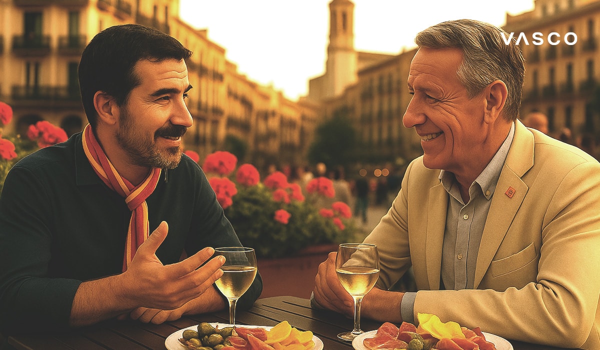 Két férfi italokat és tapast fogyaszt egy spanyol téren található szabadtéri kávézóban, beszélgetésbe merülve a virágok és az építészeti elemek hátterében.
