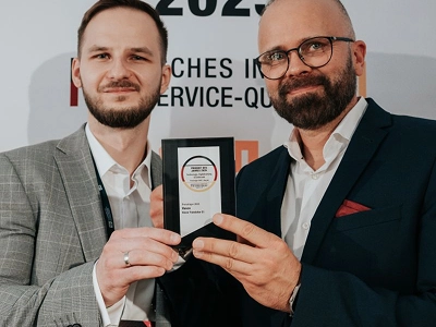 Egy széles látószögű fénykép a díjátadó színpadát ábrázolja az ülő közönség háta mögül. A színpad bal oldalán egy sötét öltönyös férfi és egy hosszú fehér ruhás nő áll, kezükben táblagépekkel, a közönség felé fordulva. A sötét háttérrel rendelkező színpad közepén egy nagy képernyőn a tetején ragyogó, borostyánszínű szöveg olvasható: „WIZJA I INNOVACJE”. Alatta, a képernyő közepén egy nagy, ragyogó, rombusz alakú kristálydíj látható, alatta pedig a „Maciej Góralski” és a „Vasco Electronics” felirat. Meleg, szórt fény világítja meg a színpadot a sötét, zsúfolt nézőtérrel szemben.