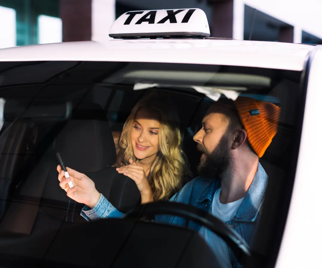 Egy férfi ül egy taxi vezetőülésében, és a hátsó ülésen ülő nőnek mutat valamit a kezében tartott tolmácson. A nő érdeklődve mosolyog. Az autó tetején egy taxitábla látható.