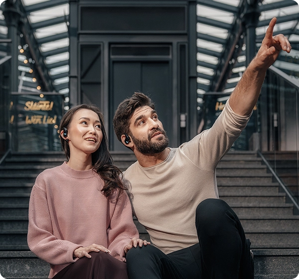 Egy férfi és egy nő egy üvegtetős, modern építészeti alkotás belsejében, betonlépcsőn ülnek. A férfi felmutat a kezével, miközben mindketten fekete Vasco Translation E1 fülhallgatót viselnek, és a férfi mozdulatának irányába néznek. 