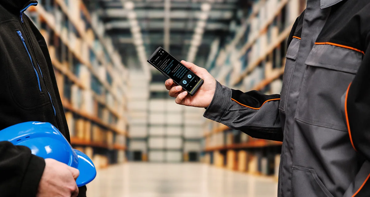 Két ember áll egymással szemben; az egyik sisakot, a másik egy Vasco Translator V4-et tart. A készülék képernyőjén egy aktív beszédfordító felület látható.