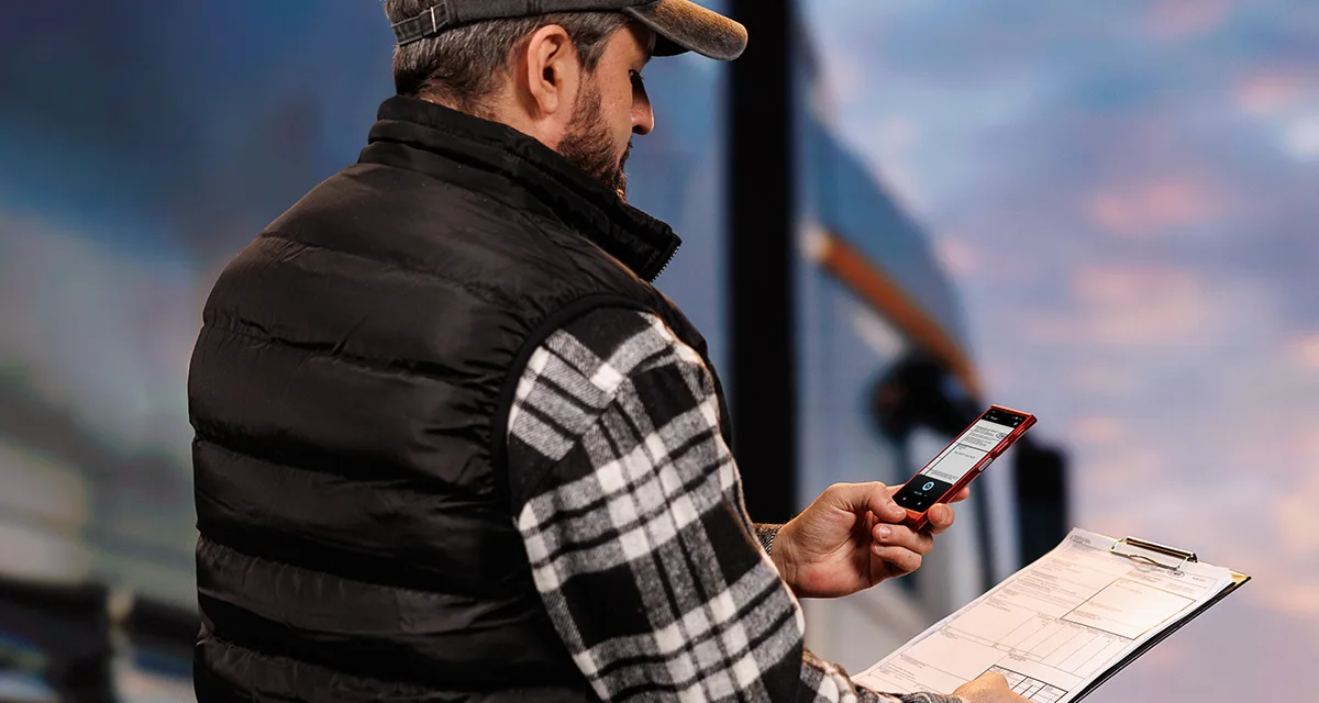 Kamionsofőr áll egy teherautó előtt, egyik kezében dokumentumokat, a másikban egy Vasco Translator V4-et tart. A készülék képernyőjén a fotó fordítás funkció látható.