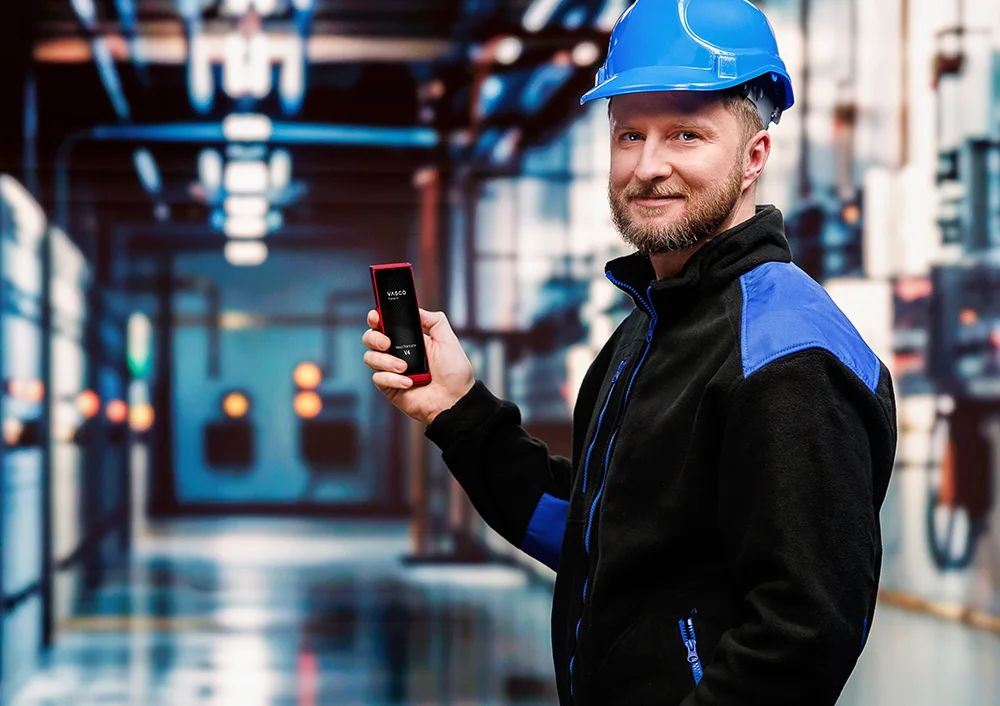 Kamionsofőr a teherautója előtt, egyik kezében papírokat, a másikban egy Vasco Translator V4-et tart, a készülék képernyőjén a fotófordítási funkció látható.