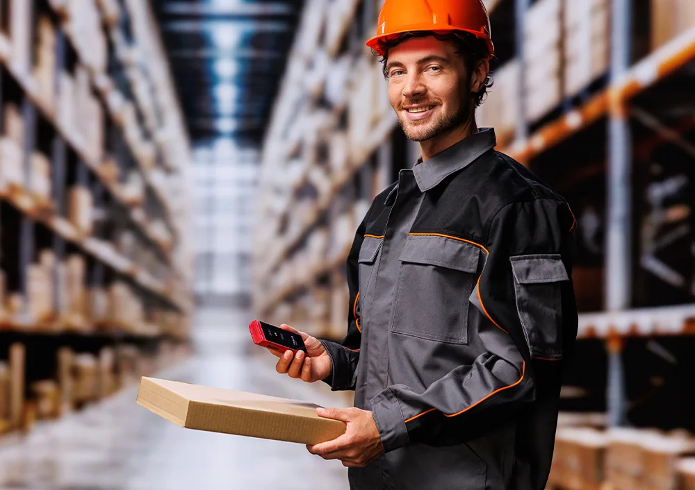 Raktári munkás munkaruhában és sisakban, egyik kezében egy Vasco Translator Q1-et, a másikban egy csomagot tart, és a csomagon található dokumentumokat/címkéket mutatja.