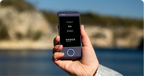 Közelkép egy kézről, amely egy lila Vasco elektronikus fordítógépet tart a kezében, világos kültéri háttér előtt. A képernyőn a „Hello!”, „¡Hola!” üdvözlések és a „VASCO Translator” logó látható. A háttérben egy napfényes tengerparti táj látható lágy fókuszú felvételen: kék víz és világos színű sziklás sziklák tiszta égbolt alatt. A készüléket tartó személy világosszürke vagy ezüstszínű, texturált kabátujjat visel. 