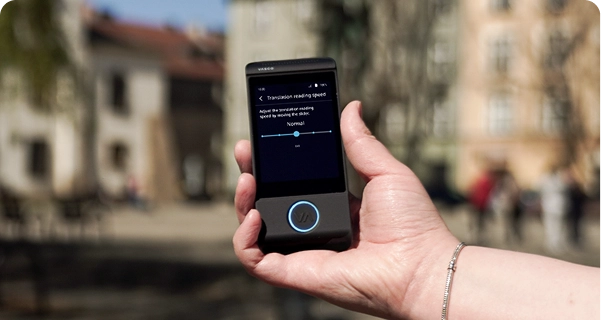 Közelkép egy ember kezéről, amely egy fekete Vasco elektronikus fordítót tart, háttérben egy történelmi épületekkel tarkított városi tér elmosódott külső képe látható. A készülék képernyőjén a „Fordítás olvasási sebessége” beállítások jelennek meg: egy „Normál” állásba állított csúszka és egy megjegyzés, amely így szól: „A csúszka mozgatásával állítsa be a fordítás olvasási sebességét.” A készüléket tartó személy ezüst karkötőt visel. A ragyogó napfény kiemeli a képernyő felületét, így a beállítások jól láthatók. 