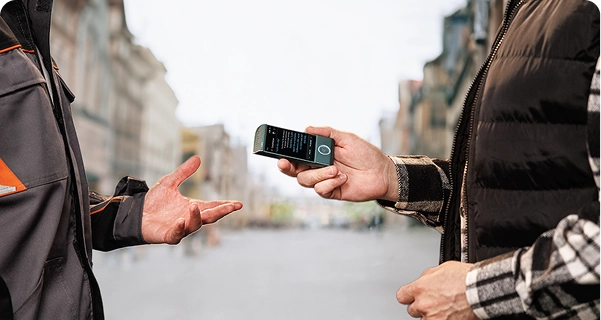 Egy férfi, aki kockás ing felett fekete, steppelt mellényt visel, vízszintesen tartja a türkizkék Vasco elektronikus fordítót egy másik ember felé egy városi utcán. A második személy, aki szürke-narancssárga munkakabátot visel, kinyújtja a kezét, mintha éppen beszélgetnének. A készülék képernyőjén egy szöveges fordítófelület látható. A háttérben egy széles, elmosódott kilátás nyílik egy európai stílusú utcára, magas épületekkel, ragyogó, felhős ég alatt. 